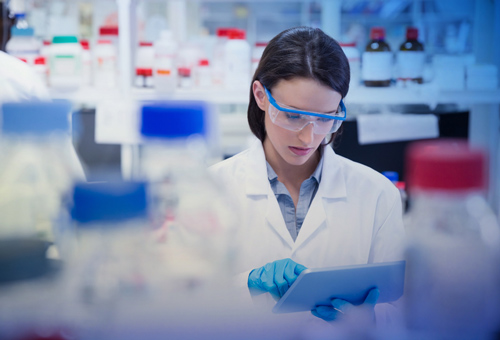 A woman in lab coat and goggles looking at tablet.
